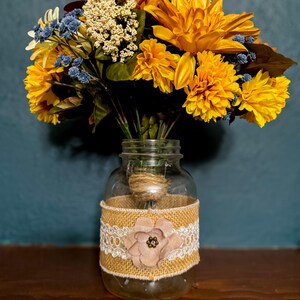 May include: A bouquet of yellow and blue artificial flowers in a clear glass jar. The jar is decorated with a burlap and lace band, and a pink fabric flower. The arrangement sits on a wooden surface.