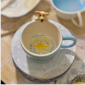 May include: A ceramic mug with a light blue handle and rim, featuring a corgi dog figurine on the rim. The interior has a yellow starburst design with blue and yellow accents. The mug has the text "Every day is be..." on the side.