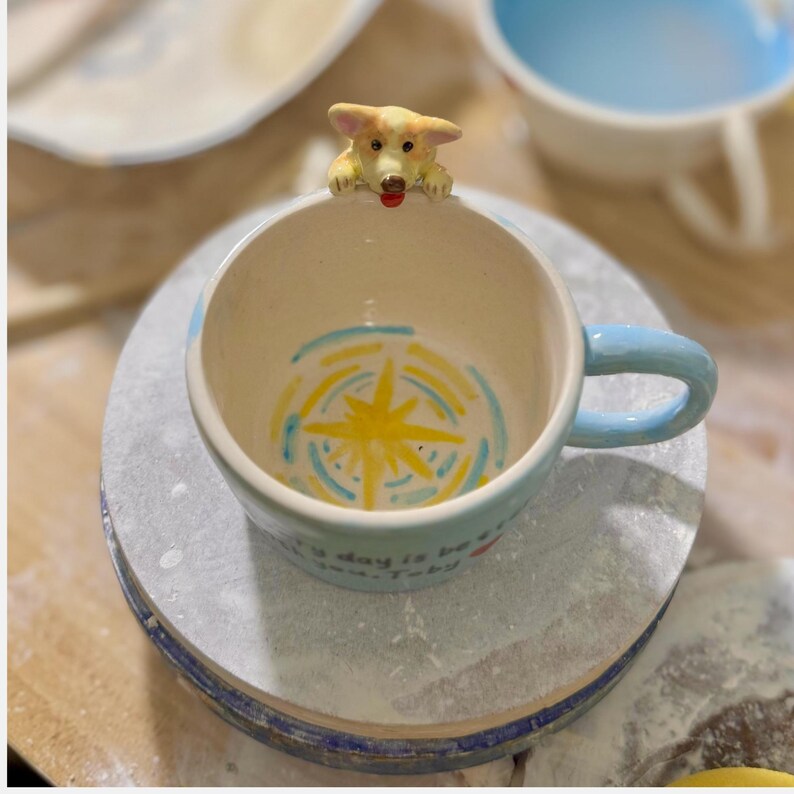 May include: A ceramic mug with a light blue handle and rim, featuring a corgi dog figurine on the rim. The interior has a yellow starburst design with blue and yellow accents. The mug has the text "Every day is be..." on the side.