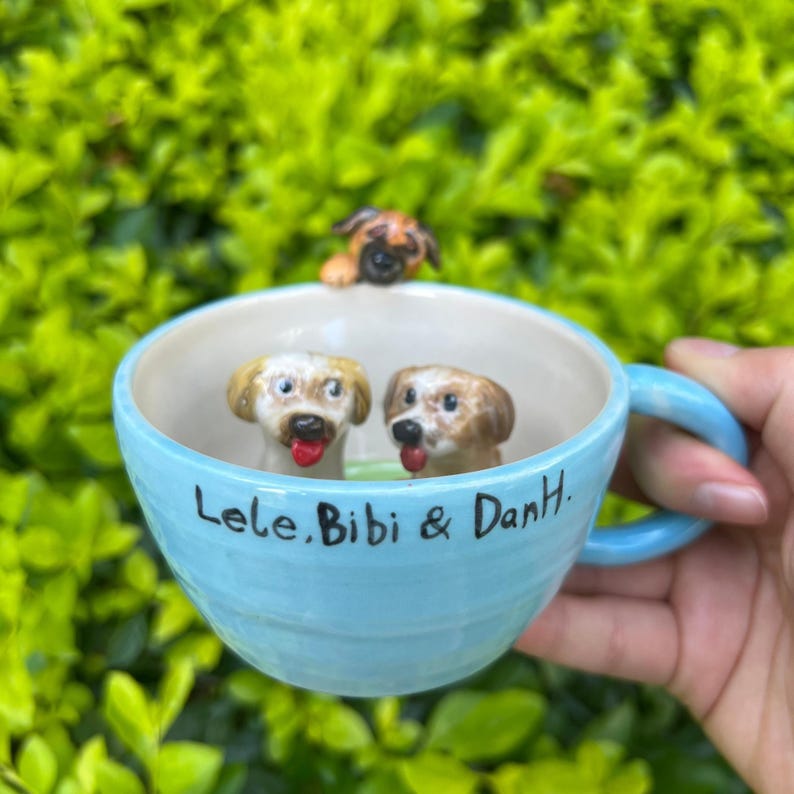 May include: A light blue ceramic mug with three dog figurines inside. The mug has a light blue handle and the names "Lele, Bibi & DanH." are written on the side. The background is a blurred green.