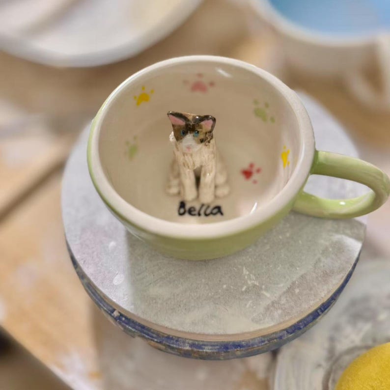 May include: A handmade ceramic mug with a light green handle and rim. Inside, a small cat figurine sits, with the name "Bella" written below. The mug's interior features paw prints and star designs.