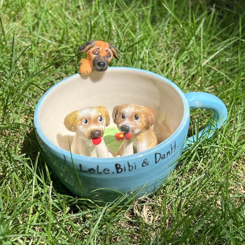 May include: A light blue ceramic teacup with three dog figurines. Two dogs are inside the cup, and one is peeking over the rim. The cup has the names "Lele, Bibi & DanH" written on it. The teacup is sitting in green grass.