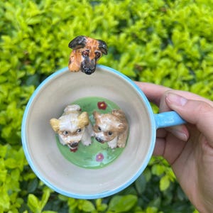 May include: A ceramic mug with a light blue handle and rim, featuring three dog figurines. Two small dogs are inside the mug on a green base with red flowers, and a larger dog head is on the rim.