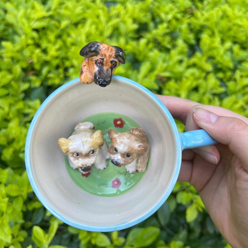 May include: A ceramic mug with a light blue handle and rim, featuring three dog figurines. Two small dogs are inside the mug on a green base with red flowers, and a larger dog head is on the rim.