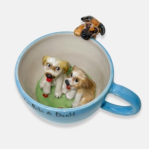 May include: A ceramic teacup with a light blue handle and rim, featuring three puppy figurines. Two puppies sit inside the cup on a green base, while a third puppy peeks over the rim. The cup is inscribed with the text "Bibi & DanH."