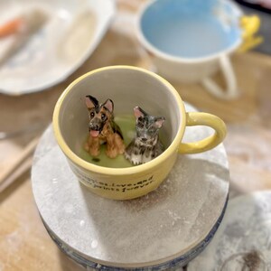 May include: A yellow ceramic mug with a handle, featuring two small dog figurines inside. One is a German Shepherd, the other a gray and black terrier. The mug has the text "memories, love, and prints forever" on the side.
