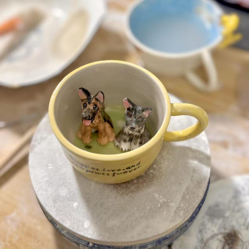 May include: A yellow ceramic mug with a handle, featuring two small dog figurines inside. One is a German Shepherd, the other a gray and black terrier. The mug has the text "memories, love, and prints forever" on the side.