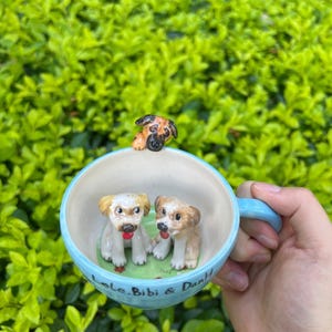May include: A light blue ceramic mug with three dog figurines. Two puppies sit inside the mug on a green base, and one puppy is perched on the rim. The mug has the text "Lele, Bibi & DanH".