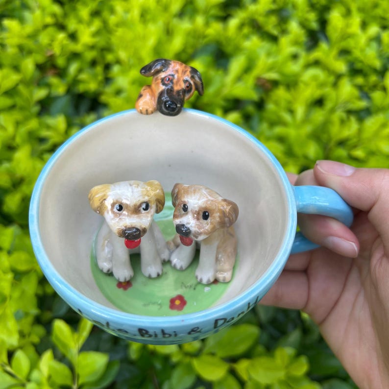 May include: A light blue ceramic mug with three dog figurines. Two puppies sit inside the mug on a green base with red flowers, and a third dog is perched on the rim. The mug has the names "Bibi & Dank" written on the side.