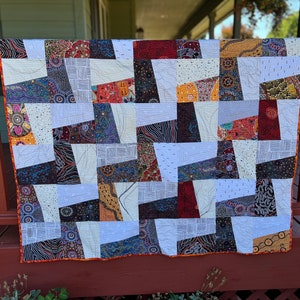 May include: A handmade quilt with a patchwork design. The quilt features a variety of patterned fabrics in shades of white, brown, orange, and blue. The quilt is displayed on a wooden railing.