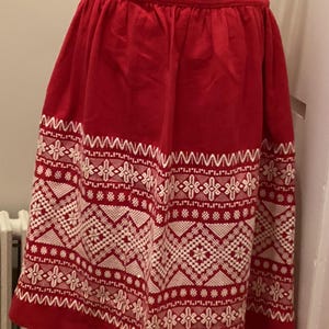May include: A red skirt featuring a wide band of white geometric embroidery near the hem. The pattern includes repeating diamond shapes, star-like motifs, and zigzag lines. The skirt has a gathered waistband and a flared, knee-length silhouette.