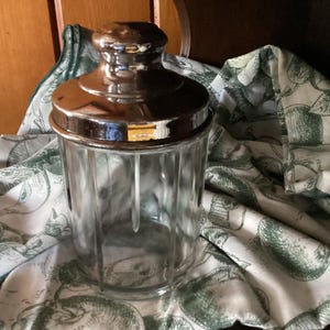 May include: A clear glass canister with vertical fluted sides and a polished metallic lid featuring a rounded knob handle. The container is cylindrical in shape and sits on a white fabric surface decorated with a green printed fruit pattern.