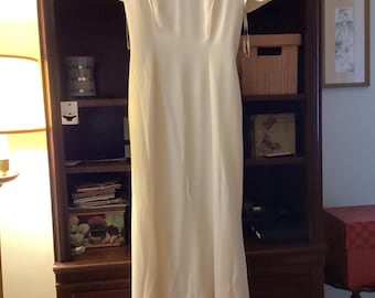 May include: Ivory-colored formal gown with short sleeves and a scoop neckline. The dress is floor-length and hangs on a wooden hanger. The dress is made of a smooth, flowing fabric. The dress is hanging in front of a dark wooden bookcase.