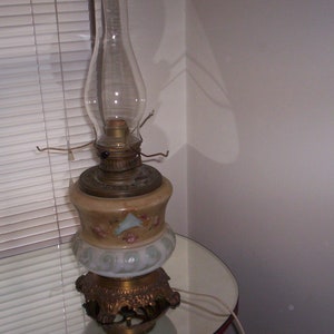 May include: A vintage oil lamp with a white and gold base and a clear glass chimney. The lamp has a floral design on the base.