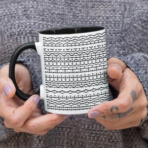 Puede incluir: Una taza de cerámica blanca con interior y asa negros. La taza está decorada con un patrón negro repetido de varias formas geométricas. La taza es sostenida por una persona que lleva un suéter gris.
