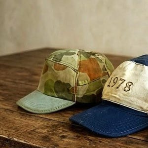 May include: Three baseball caps featuring varied designs. The left cap has a green camouflage pattern with a light blue brim. The centre cap is navy blue with a white front panel displaying the text 1978. The right cap features camouflage fabric, a triangular patch, and a small American flag patch.
