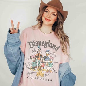 Puede incluir: Una camiseta rosa claro de Disneyland con personajes cl&aacute;sicos de Disney como Mickey Mouse, Minnie Mouse y Donald Duck. La camiseta tiene el texto "Disneyland California" y "EST. 1955". La persona lleva un sombrero marr&oacute;n y una chaqueta vaquera.