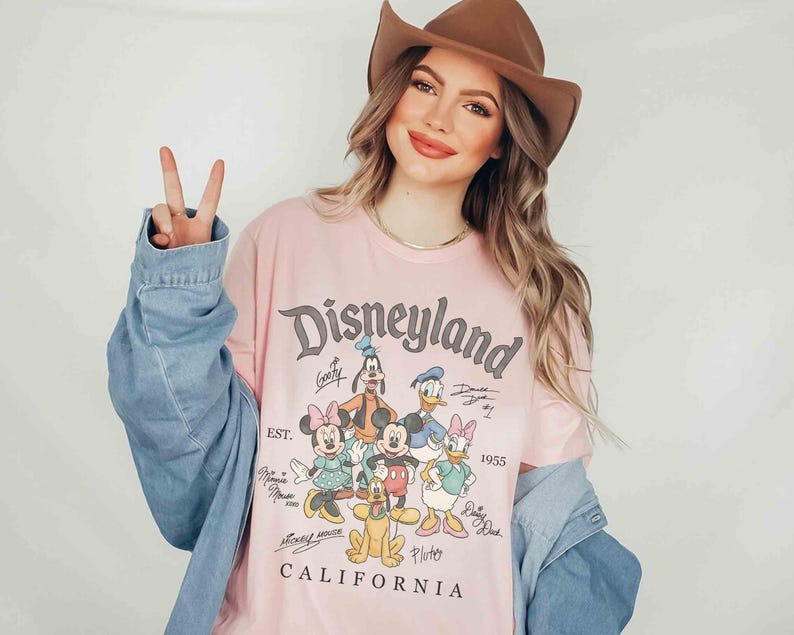 Puede incluir: Una camiseta rosa claro de Disneyland con personajes cl&aacute;sicos de Disney como Mickey Mouse, Minnie Mouse y Donald Duck. La camiseta tiene el texto "Disneyland California" y "EST. 1955". La persona lleva un sombrero marr&oacute;n y una chaqueta vaquera.