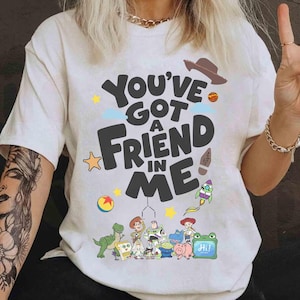 K&ouml;nnte beinhalten: Cremefarbenes T-Shirt mit dem Schriftzug "You've Got a Friend in Me" und Cartoon-Charakteren. Das Design umfasst einen Cowboyhut, eine Rakete und andere verspielte Elemente. Das Shirt ist ein Freizeitartikel.