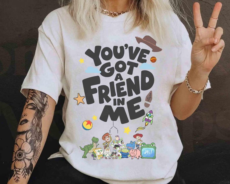 K&ouml;nnte beinhalten: Cremefarbenes T-Shirt mit dem Schriftzug "You've Got a Friend in Me" und Cartoon-Charakteren. Das Design umfasst einen Cowboyhut, eine Rakete und andere verspielte Elemente. Das Shirt ist ein Freizeitartikel.