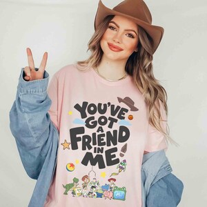 K&ouml;nnte beinhalten: Hellrosa T-Shirt mit dem Text "You've Got a Friend in Me" in Schwarz, mit Cartoon-Grafiken. Das Shirt zeigt einen Cowboyhut, einen Stern, einen Ball und andere Charakterillustrationen. Teil einer Bekleidungskollektion.