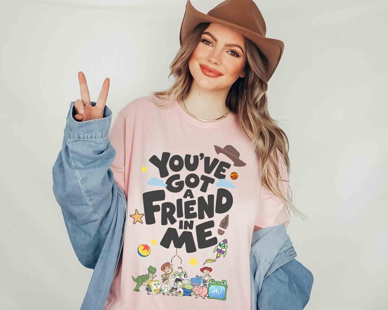 K&ouml;nnte beinhalten: Hellrosa T-Shirt mit dem Text "You've Got a Friend in Me" in Schwarz, mit Cartoon-Grafiken. Das Shirt zeigt einen Cowboyhut, einen Stern, einen Ball und andere Charakterillustrationen. Teil einer Bekleidungskollektion.