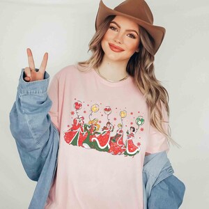 May include: A light pink long-sleeve t-shirt featuring a graphic of cartoon characters in red and green dresses holding balloons. The balloons are red, yellow, and green. The shirt is worn with a brown cowboy hat and a denim jacket.