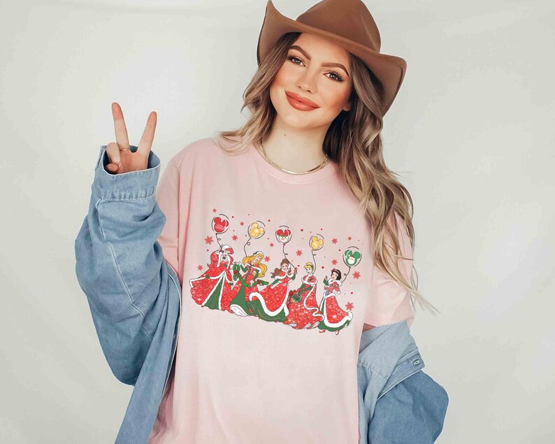 May include: A light pink long-sleeve t-shirt featuring a graphic of cartoon characters in red and green dresses holding balloons. The balloons are red, yellow, and green. The shirt is worn with a brown cowboy hat and a denim jacket.