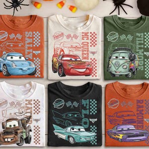 May include: Six folded t-shirts featuring cartoon car designs. The shirts are in various colors, including orange, white, green, and black. Each shirt displays a different car character with the names "Sally", "McQueen", "Fillmore", "Mater", and "Ramone".