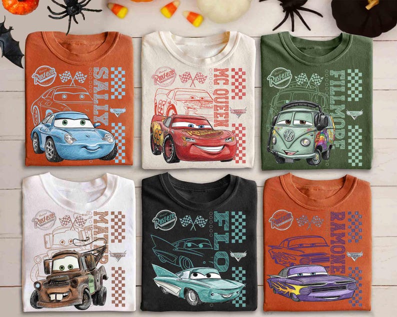 May include: Six folded t-shirts featuring cartoon car designs. The shirts are in various colors, including orange, white, green, and black. Each shirt displays a different car character with the names "Sally", "McQueen", "Fillmore", "Mater", and "Ramone".