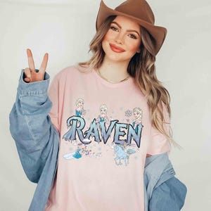 Puede incluir: Una camiseta rosa claro con el nombre "Raven" en una fuente azul hielo y ilustraciones de personajes animados. La camiseta es de cuello redondo.