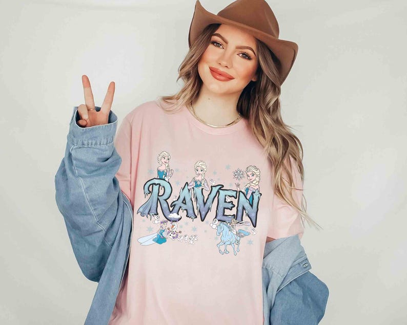 Puede incluir: Una camiseta rosa claro con el nombre "Raven" en una fuente azul hielo y ilustraciones de personajes animados. La camiseta es de cuello redondo.