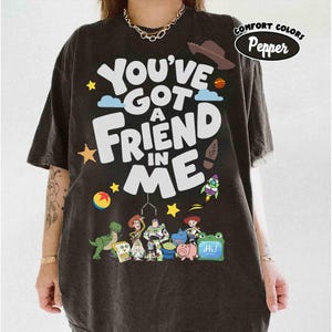 K&ouml;nnte beinhalten: Dunkelgraues T-Shirt mit dem Schriftzug "You've Got A Friend In Me" in gro&szlig;en wei&szlig;en Buchstaben. Das Shirt zeigt Zeichentrickfiguren und Illustrationen eines Cowboyhuts, einer Rakete und anderer verspielter Elemente.