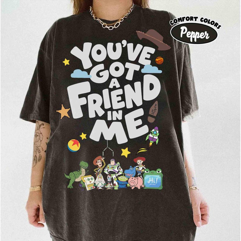 K&ouml;nnte beinhalten: Dunkelgraues T-Shirt mit dem Schriftzug "You've Got A Friend In Me" in gro&szlig;en wei&szlig;en Buchstaben. Das Shirt zeigt Zeichentrickfiguren und Illustrationen eines Cowboyhuts, einer Rakete und anderer verspielter Elemente.