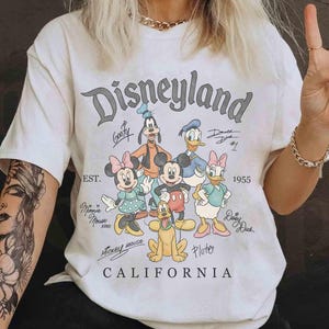 Puede incluir: Camiseta crema de Disneyland California con un gr&aacute;fico de Mickey Mouse, Minnie Mouse, Donald Duck, Daisy Duck, Goofy y Pluto. La camiseta tiene el texto "Disneyland" y "1955" impreso.
