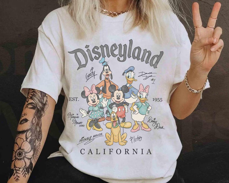 Puede incluir: Camiseta crema de Disneyland California con un gr&aacute;fico de Mickey Mouse, Minnie Mouse, Donald Duck, Daisy Duck, Goofy y Pluto. La camiseta tiene el texto "Disneyland" y "1955" impreso.