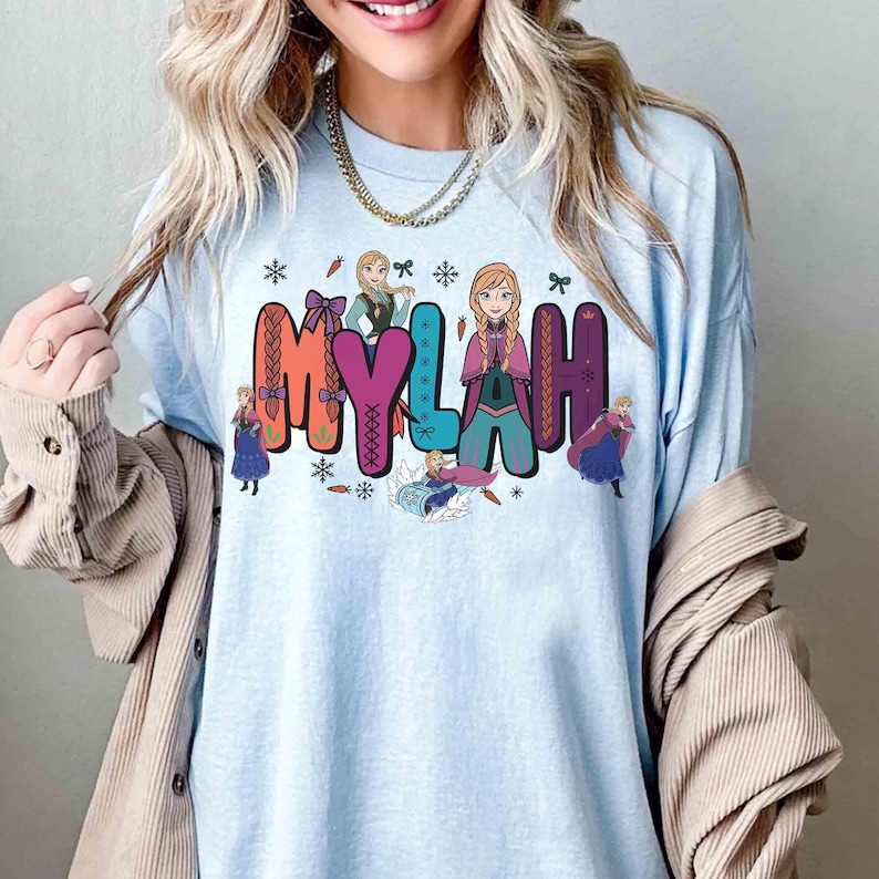 Puede incluir: Camiseta azul claro con el nombre "MYLAH" en letras de bloque coloridas, decorada con copos de nieve, lazos y personajes de dibujos animados. Las letras son de color naranja, rosa, verde y morado.