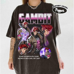 May include: Dark gray t-shirt featuring the word "GAMBIT" in a gradient pink and white font. The shirt displays multiple images of a character with red hair and a purple and brown outfit. Text on the shirt reads "PLAYIN' FOR KEEPS IS STILL PLAYIN', MON AMI, SO TAKE A CARD...ANY CARD!"