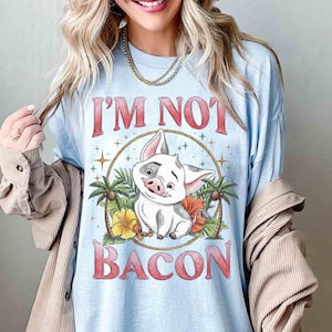Puede incluir: Camiseta azul claro con el texto rojo "I'M NOT BACON". El dise&ntilde;o presenta un cerdo de dibujos animados sonriente, rodeado de flores tropicales y palmeras. El cerdo tiene manchas blancas y negras.