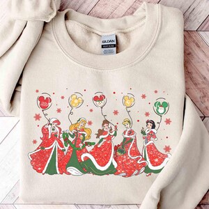 May include: Beige sweatshirt featuring a festive design with Disney princesses in red and green holiday dresses. Each princess holds a balloon with a Mickey Mouse head design. Snowflakes and Mickey balloons complete the holiday theme.