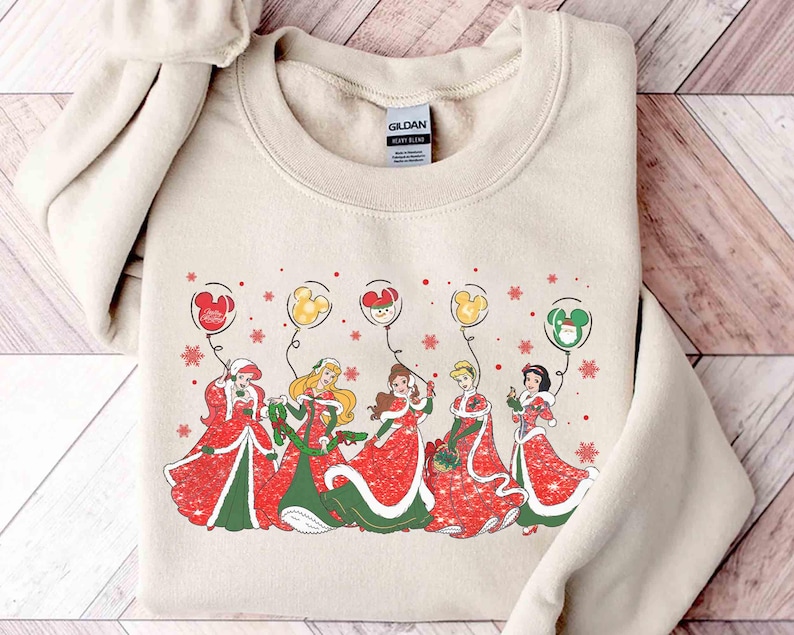 May include: Beige sweatshirt featuring a festive design with Disney princesses in red and green holiday dresses. Each princess holds a balloon with a Mickey Mouse head design. Snowflakes and Mickey balloons complete the holiday theme.