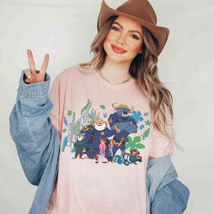 May include: Pale pink long-sleeved t-shirt with a vibrant cartoon graphic. The design includes a blue bull-like character, a tiger, and other figures. The shirt is a light pink colour and has long sleeves.