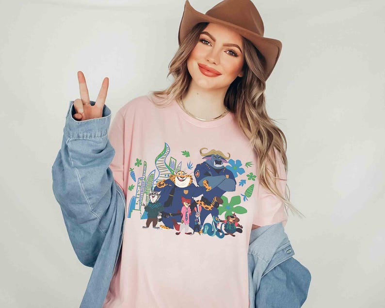 May include: Pale pink long-sleeved t-shirt with a vibrant cartoon graphic. The design includes a blue bull-like character, a tiger, and other figures. The shirt is a light pink colour and has long sleeves.