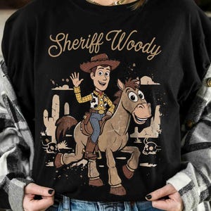 May include: Black t-shirt featuring a cartoon illustration of Sheriff Woody riding a horse. The design includes the text "Sheriff Woody" in a script font, along with desert scenery and a skull. The character is waving.