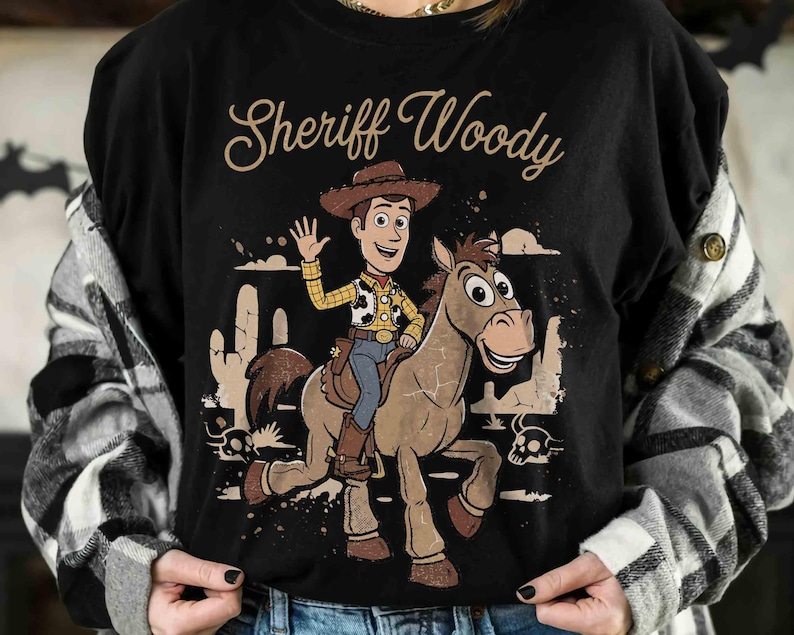 May include: Black t-shirt featuring a cartoon illustration of Sheriff Woody riding a horse. The design includes the text "Sheriff Woody" in a script font, along with desert scenery and a skull. The character is waving.