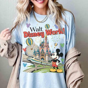 Puede incluir: Camiseta azul claro con un dise&ntilde;o de Walt Disney World. El dise&ntilde;o incluye un castillo, Mickey Mouse, un monorra&iacute;l y el texto "Walt Disney World" en rojo y amarillo. Globos y fuegos artificiales completan el dise&ntilde;o.
