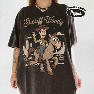 May include: Dark gray t-shirt featuring a cartoon illustration of Sheriff Woody riding a horse. The shirt has the words "Sheriff Woody" in a stylized font. The shirt also has the words "Comfort Colors Pepper" in a circular design.
