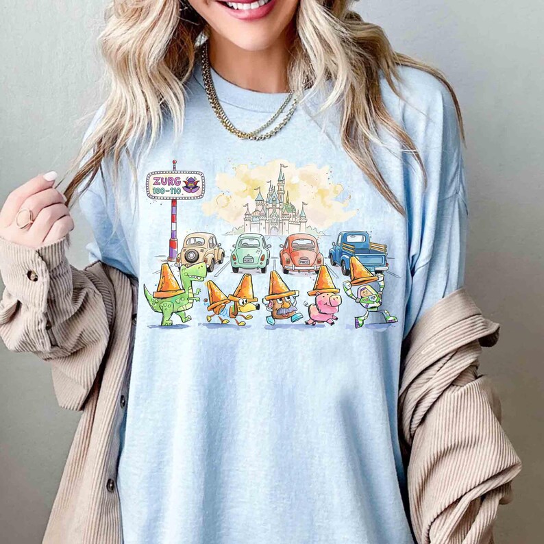 May include: Pale blue long-sleeved shirt with a colourful cartoon design. The graphic shows cartoon characters wearing orange traffic cones, vintage cars, a castle, and the text "Zurg 100-110". The shirt has a loose fit.