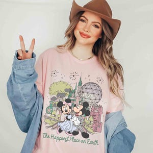 May include: Pink long-sleeve t-shirt featuring a graphic of Mickey and Minnie Mouse in front of a castle and a geodesic sphere. The text "The Happiest Place on Earth" is printed below the graphic. The shirt is worn with a denim jacket and a brown hat.