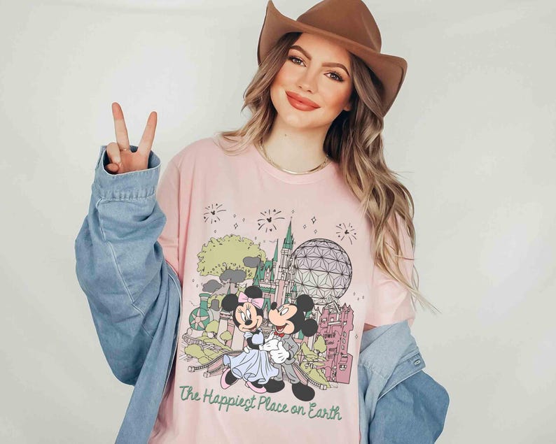 May include: Pink long-sleeve t-shirt featuring a graphic of Mickey and Minnie Mouse in front of a castle and a geodesic sphere. The text "The Happiest Place on Earth" is printed below the graphic. The shirt is worn with a denim jacket and a brown hat.